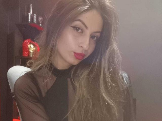LeilaGoddes ha lunghi capelli castani ondulati e rossetto rosso, ed indossa una maglia nera trasparente. È leggermente inclinata in avanti, e guarda la fotocamera in modo seducente. Candele rosse sullo sfondo..