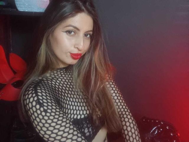 LeilaGoddes, een aantrekkelijke vrouw met lang bruin haar, rode lippenstift en een zwarte kousenbroektop, kijkt verleidelijk. schouder. Rood licht op de achtergrond creëert een sensuele sfeer..