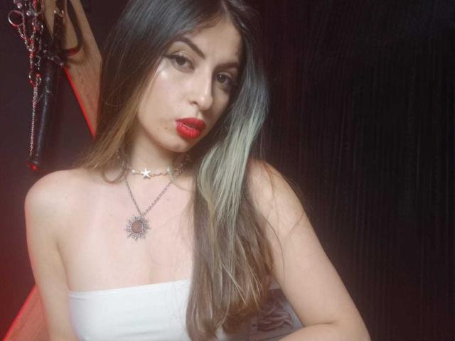 Foto van LeilaGoddes, met een medium bruine huid, lang zwart haar met groene highlights, rode lipstick, een wit topje zonder bandjes. Zilveren ster ketting, sensuele blik, subtiele BDSM elementen, donkere achtergrond, zelfverzekerde pose..