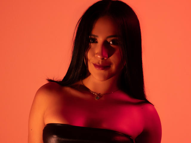 Photograph of AnyaJay with long black hair, wearing a strapless black leather top, illuminated by a vivid red light. Her dark eyes and lips are highlighted, creating a seductive, intense expression.
