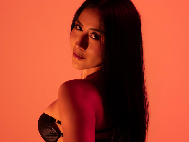 Fotografía de AnyaJay con cabello negro largo, mirando por encima del hombro, vistiendo un sujetador de encaje negro. Fondo rojo brillante, iluminación intensa, sombras en su rostro, un ambiente sexy y seductor..