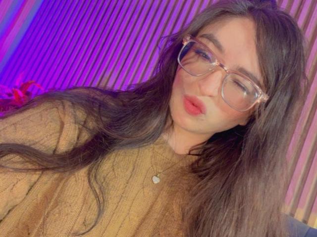 Eva McAllister, che indossa occhiali grandi e un maglione marrone, ha lunghi capelli neri e labbra rosse. La sua espressione è leggermente seducente. Sfondo neon viola e blu. Leggera tensione sessuale.