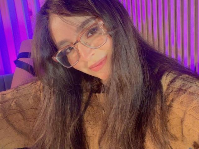 Close-up of EvaMcAllister with long black hair, wearing clear glasses and a brown patterned sweater. She leans forward, smiling seductively, with purple and blue lighting in the background. Fucking sexy.