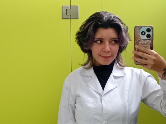 DarckVelvet maakt een selfie met een smartphone in een heldergele kamer. Ze dragen een witte laboratoriumjas, hebben rommelig donker haar en een subtiele glimlach. Een hand die een telefoon vasthoudt is zichtbaar..