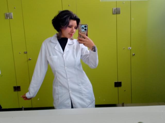 DarckVelvet mit kurzen, lockigen schwarzen Haaren macht ein Selfie vor einem Spiegel in einer Umkleidekabine. In einem engen, weißen Laborkittel lehnen sie sich vor und betonen dabei ihre weiblichen Formen und ihre kleinen Brüste. Ein Telefon mit einer blauen Hülle halten..