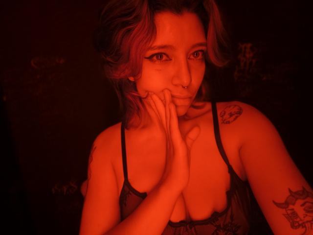 DarckVelvet with shoulder-length, wavy hair, red-lit, gazing at camera, hands near face, black tank top, visible tattoos on arm, intense expression, sexy, red glow, seductive pose, dark background.