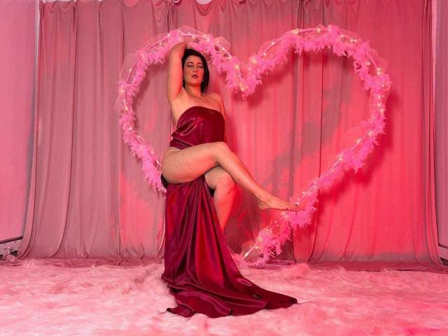 Yessi Soto está sentada com as pernas cruzadas, vestindo um vestido vermelho de cetim sem alças, apoiada em um coração rosa feito de penas. Cenário iluminado em tons de rosa. O fundo é composto por cortinas penduradas, e o chão está coberto por pele branca..