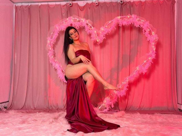 YessiSoto, uma mulher asiática atraente com longos cabelos pretos, sentada em um vestido de cetim vermelho, com as pernas cruzadas, em frente a um fundo rosa. Fundo com penas em formato de coração, iluminação vermelha, pose provocativa, olhar sedutor..
