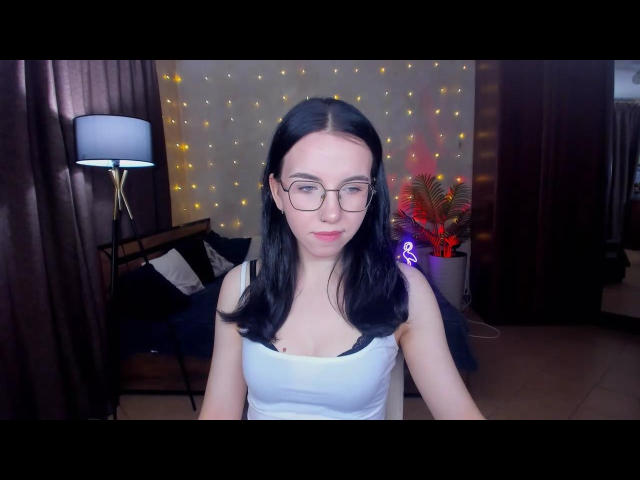 Tricia Barnes mit glattem, schwarzem Haar, die ein weißes Tanktop, eine Brille und einen neutralen Gesichtsausdruck trägt. Ein schwach beleuchteter Raum mit Lichterketten, einer Lampe und dunklen Vorhängen im Hintergrund. Leichte sexuelle Anspannung.