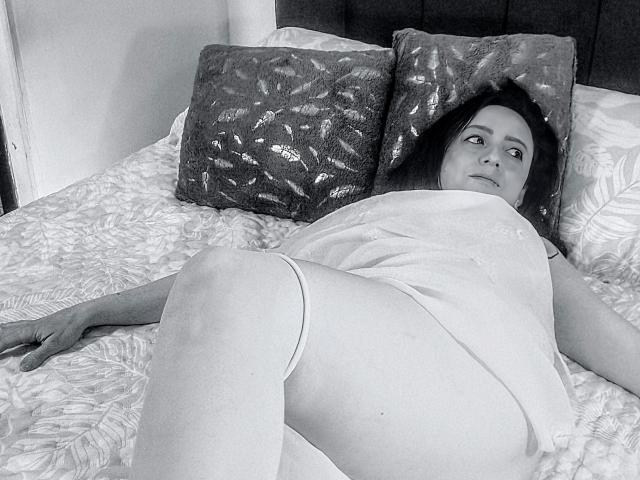 Photographie en noir et blanc de CatalinaMonyque allongée sur un lit avec des oreillers brillants, portant une chemise blanche et sans pantalon. Les jambes écartées, exposant son vagin, elle regarde vers le haut avec une expression détendue..