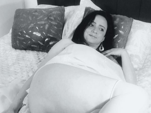 Fotografia in bianco e nero di CatalinaMonyque sdraiata su un letto, gambe divaricate, che indossa una maglietta bianca, con la pancia prominente, di colore scuro. capelli, cuscini ricamati, espressione seducente, posa informale, tensione sessuale.