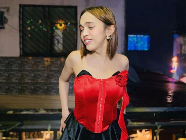 Fotografia de TinyAsian-hot, em pé, com os olhos fechados, vestindo um corpete vermelho com fita preta, saia preta e brincos. O fundo apresenta superfícies refletoras com iluminação fraca, criando uma atmosfera sensual, como a de uma boate..