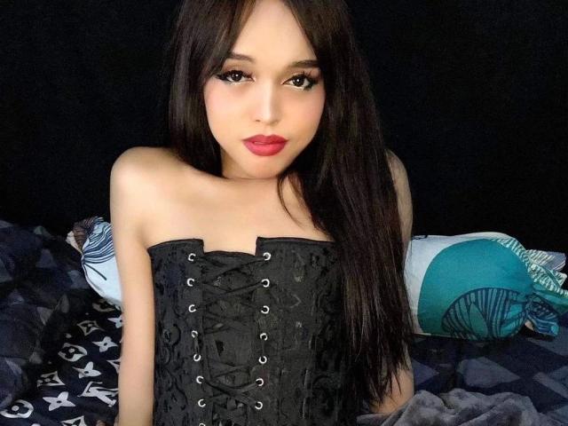 Photographie d'une jeune femme asiatique, TinyAsian-hot, avec de longs cheveux noirs, du rouge à lèvres rouge et un corset noir, assise sur une lit avec des oreillers sombres et bleu canard. Extrêmement séduisant, regardant directement l'appareil photo..