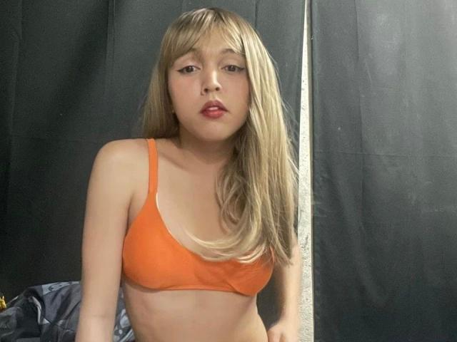 Fotografía de una joven asiática delgada, con cabello rubio largo, que lleva un sujetador naranja y posa frente a una cortina negra. Sus labios rojos están ligeramente entreabiertos, mostrando tensión sexual..