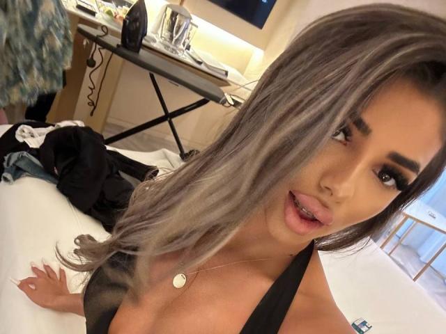 Foto em close-up de CrisstyBlair, sexy, com cabelos grisalhos longos e ondulados, lábios cheios levemente entreabertos, vestindo uma blusa preta e dourada. colar.
o fundo mostra uma mesa bagunçada com luzes. Incrivelmente atraente..