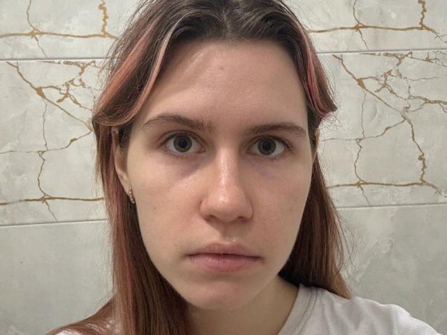 Close-up de Jeanelle Stamnos, uma jovem com pele clara, olhos castanhos e cabelo castanho liso, vestindo uma camisa branca. Fundo de parede branca rachada. Olhar inexpressivo, ligeiramente intenso..