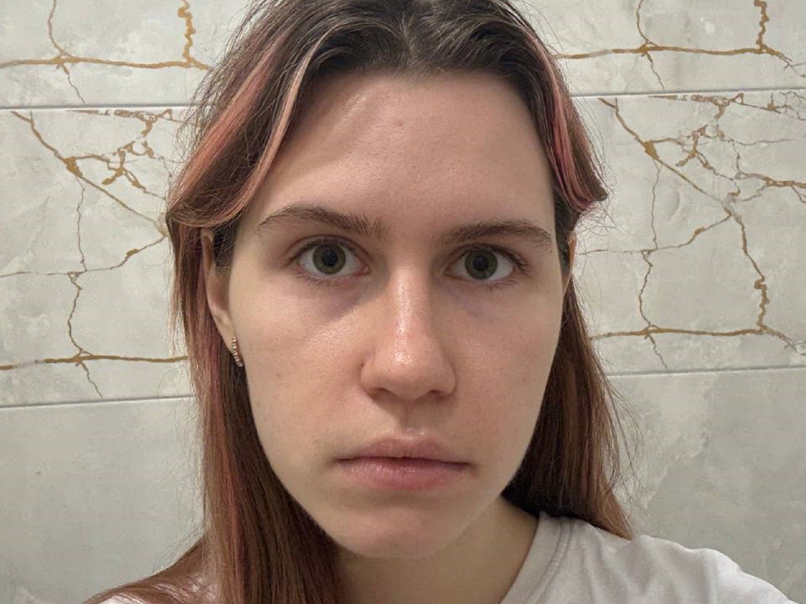 Gros plan de Jeanelle Stamnos, une jeune femme à la peau pâle, aux yeux marron et aux cheveux bruns lisses, portant une chemise blanche. Fond blanc avec un mur fissuré. Regard impassible, légèrement intense..