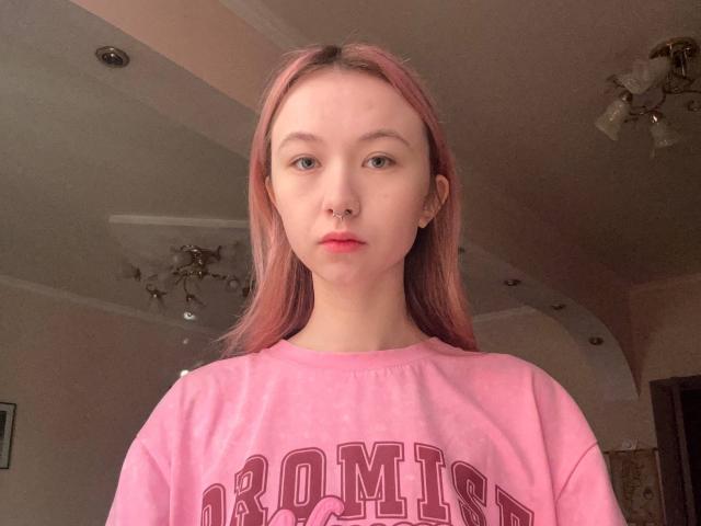 Fotografie einer jungen Frau mit heller Haut und langen, glatten, rötlich-braunen Haaren, die einen rosa Sweatshirt trägt. "ARMORY" in roten Buchstaben. Sie steht mit einem neutralen Gesichtsausdruck vor der Kamera..