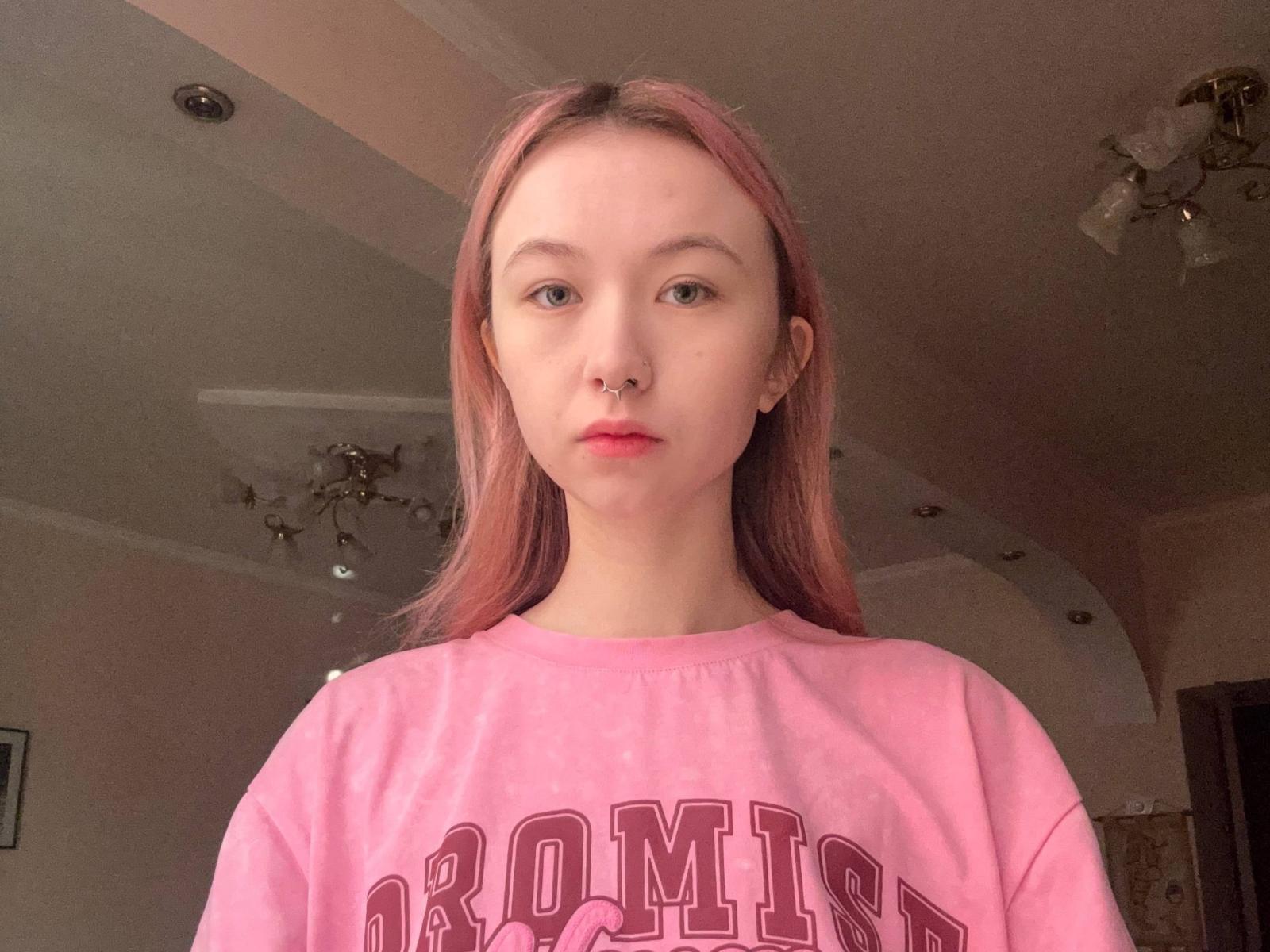 Fotografie einer jungen Frau mit heller Haut und langen, glatten, rötlich-braunen Haaren, die einen rosa Sweatshirt trägt. "ARMORY" in roten Buchstaben. Sie steht mit einem neutralen Gesichtsausdruck vor der Kamera..