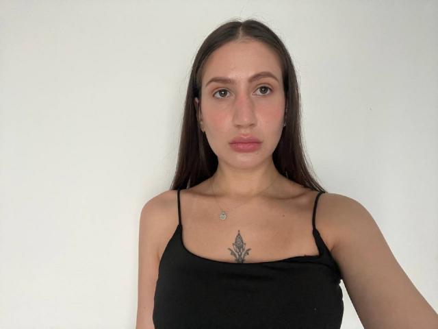 Fotografia di Fredericka Thonney con lunghi capelli scuri, che indossa una canottiera nera a spalline sottili, che mostra un piccolo tatuaggio. tra i suoi seni. Sta guardando direttamente la fotocamera con un'espressione neutra..