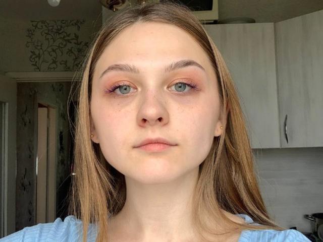 Photo en gros plan de RosannSexy, avec une peau pâle, des yeux bleus et des cheveux bruns clairs. Elle a du fard à paupières rose et une expression neutre. L'arrière-plan présente des armoires de cuisine blanches et un mur à motifs. Extrêmement sexy.