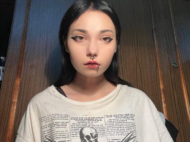 ShayHedglin has pale skin, black hair, and a nose ring. She wears a white shirt with black text and a small graphic. She looks directly at the camera with a serious expression.