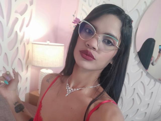 VicktoriaSunshine, una latina con cabello largo y negro, que lleva una blusa roja de encaje, gafas grandes y transparentes, y un collar brillante, tiene labios rojos y piel clara, sosteniendo sus uñas, mirando directamente a la cámara, sexy..