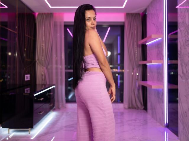 Kelany, una mujer con cabello negro largo, de pie en una habitación moderna con luces de neón. Vestía un top rosa sin tirantes y pantalones ajustados a juego. Miraba por encima del hombro, resaltando sus curvas..