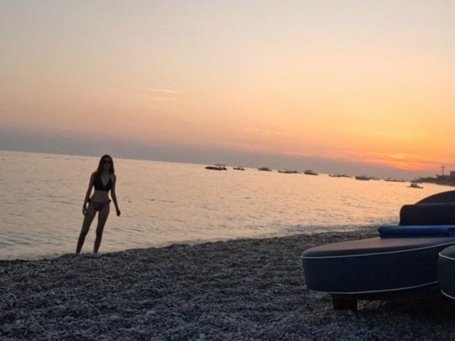 Silhouette of VictoriaPalmerfg, slender, wearing a bikini, standing on a pebbled beach at sunset, cock in bikini briefs, facing the water, two lounge chairs in the foreground, orange sky.