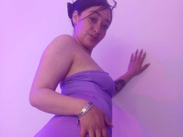 Alanna Scott, una mujer de curvas, con piel clara y cabello oscuro recogido en un moño, lleva un vestido púrpura sin tirantes y está de pie frente a pared blanca con iluminación púrpura. Mano en la pared, contorno del pene visible..