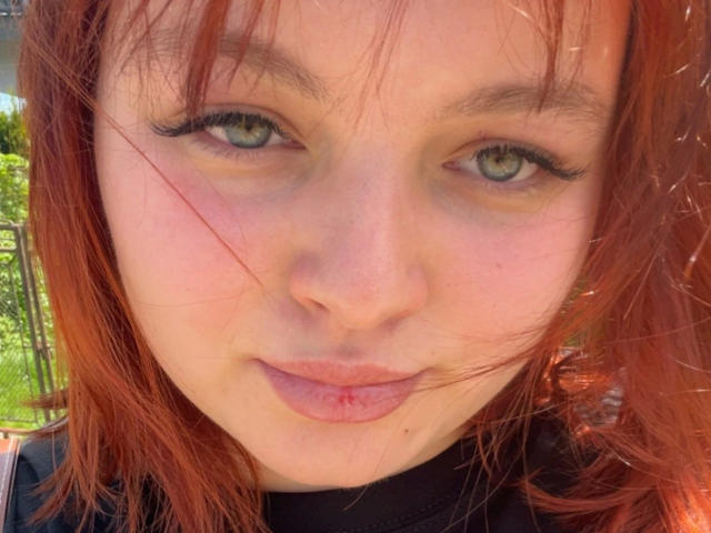 Close-up foto van MollyLolly-hot, een jonge vrouw met bleke huid, groene ogen en rood haar, die recht in de camera kijkt. met een subtiele, lichtjes open mond. Haar uitdrukking is subtiel verleidelijk..