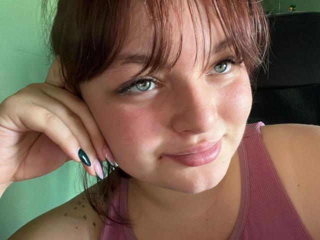 Close-up foto van Molly. Ze heeft een aantrekkelijk uiterlijk, een lichte huid, groene ogen, bruin haar en een pony. Ze draagt een roze tanktop, heeft haar hand op haar wang en heeft zwarte nagels. Met een grijns, aantrekkelijk, lichte blos..