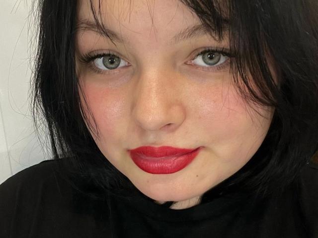Photo en gros plan de MollyLolly-hot, une femme à la peau pâle, aux cheveux noirs, aux yeux verts et au rouge à lèvres rouge vif, qui regarde fixement. regardant directement la caméra avec un léger sourire, dégageant une tension sexuelle..