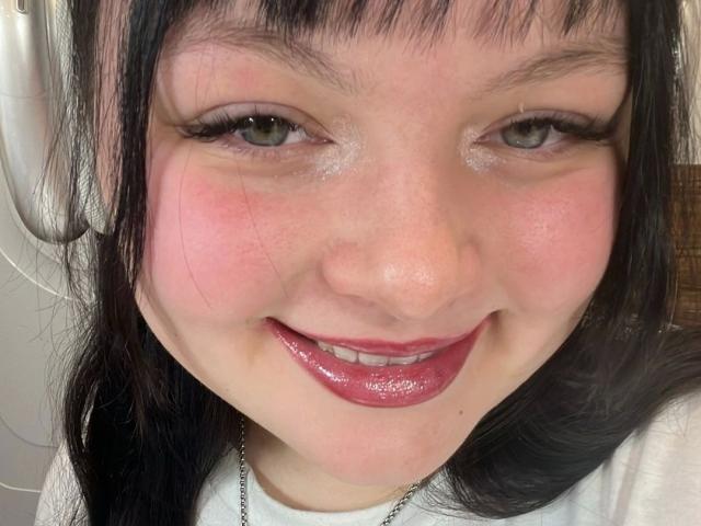 Gros plan du visage de MollyLolly-hot, avec des lèvres roses brillantes, des yeux marron foncé et une frange noire. Elle sourit, avec une légère rougeur sur ses joues. Sa peau brillante présente des pores visibles. Le coq n'est pas visible..