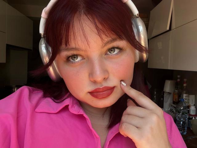Close-up foto van Molly. Ze heeft een aantrekkelijke uitstraling, een lichte huidskleur, rode lippenstift, donkerbruin haar, draagt witte koptelefoons en een roze shirt. Ze raakt haar lip aan met een vinger, en haar groene ogen kijken verleidelijk naar de camera..
