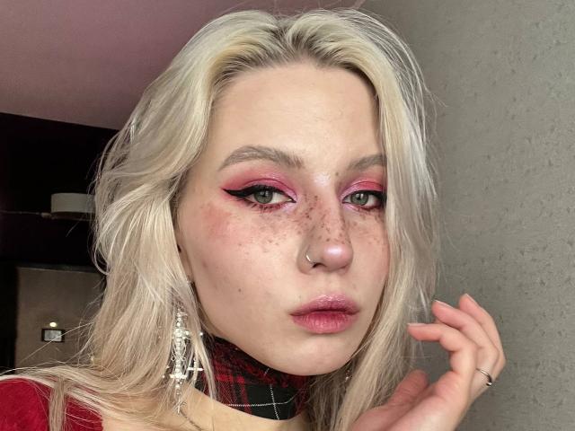 Nahaufnahme von Jacque Bridenstine mit platinblondem Haar, rosa Lidschatten und Sommersprossen. Sie trägt einen Nasenring, eine schwarze Halskette, ein rotes Oberteil und hängende Ohrringe. Hand in der Nähe des Gesichts, verführerischer Ausdruck..