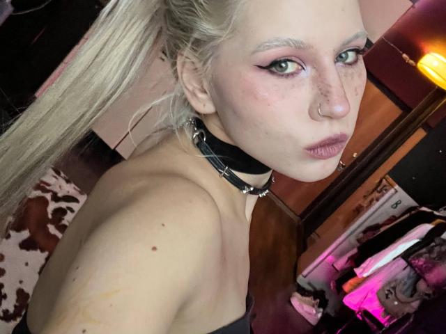 Close-up of JacqueBridenstine, a blonde woman with heavy eyeliner, nose piercing, and black choker, looking seductively at the camera, topless, with a hint of pink neon light in the background.