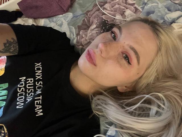 Jacque Bridestine ligt op een bed, op haar rug, met blond haar en sproeten, gekleed in een zwart shirt met witte tekst, en kijkt naar... camera, subtiele seksuele spanning, zichtbare tatoeage op de rechterarm.