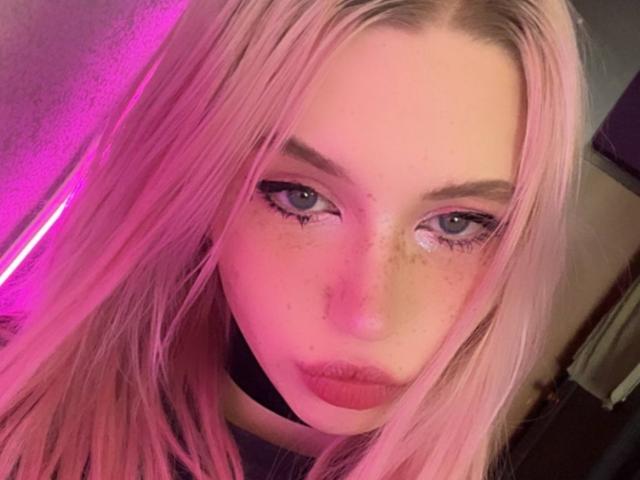 Autofoto de primer plano de Jacque Bridenstine con cabello rosa, ojos azules y pecas. La iluminación rosa resalta su rostro, creando un brillo seductor. Ella mira directamente a la cámara con una expresión seductora..