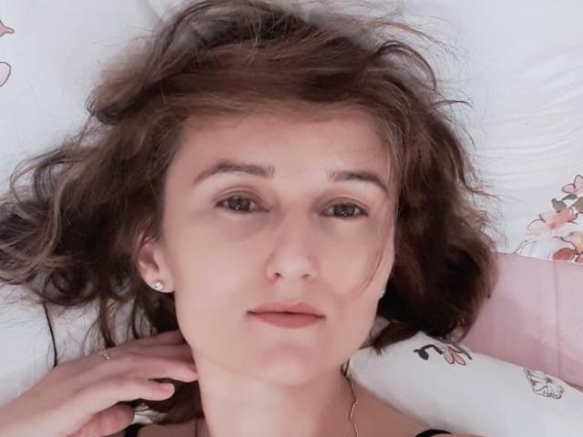 Close-up de LamourCestMoi, uma jovem com cabelos castanhos desalinhados, pele clara e olhos castanhos, deitada em uma cama branca. lençóis e almofadas com estampas florais. A mão dele toca o pescoço dela. Uma expressão incrivelmente relaxada..