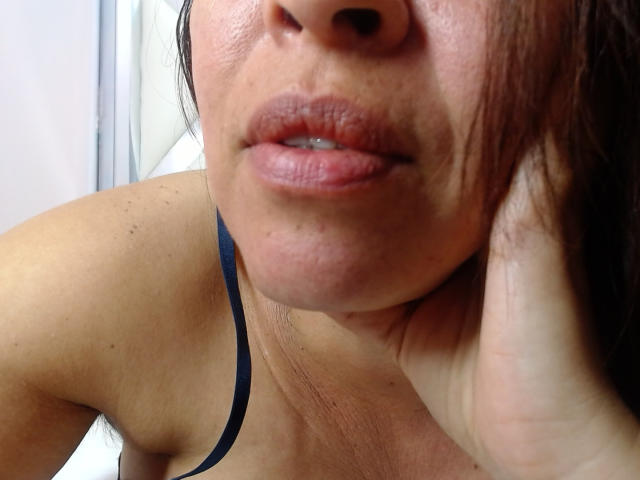 Fotografía de primer plano de LizethBells, enfocándose en su rostro y parte superior del pecho. Está ligeramente girada, con los labios entreabiertos y una correa negra visible. Su piel está sudorosa y enrojecida. Su mano descansa sobre su cuello..