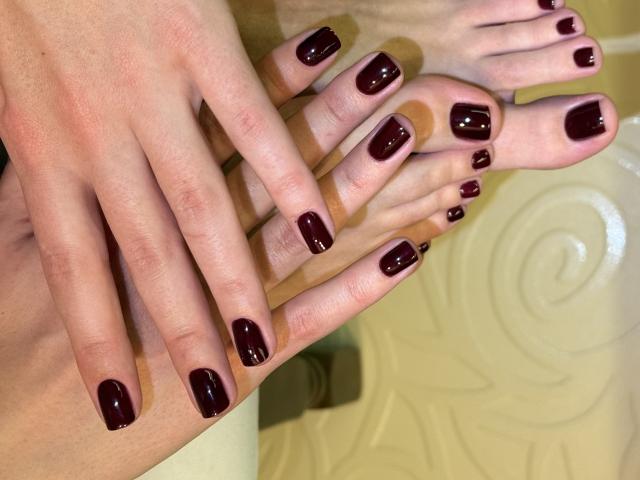 Close-up dos pés e das mãos de MargoFun, com esmalte roxo escuro brilhante. Pés cruzados nos tornozelos, mãos apoiadas em cima. A pele é lisa, levemente brilhante. O fundo está desfocado..