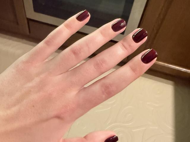 Close-up of a pale hand with dark red, glossy nail polish, palm facing up, fingers slightly spread. Background includes blurred kitchen cabinet and oven. Shadows create contrast. Fingers look like they're ready to touch something.