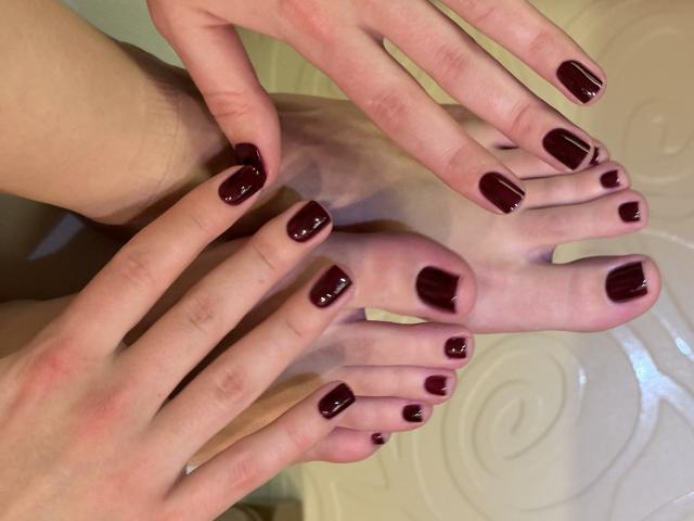 Close-up das mãos e pés de MargoFun, com os dedos espalhados sobre os pés, ambos com esmalte vermelho escuro brilhante. Pele pálida, sombras que realçam as curvas. Tensão erótica, dedos tocando os pés de forma íntima..