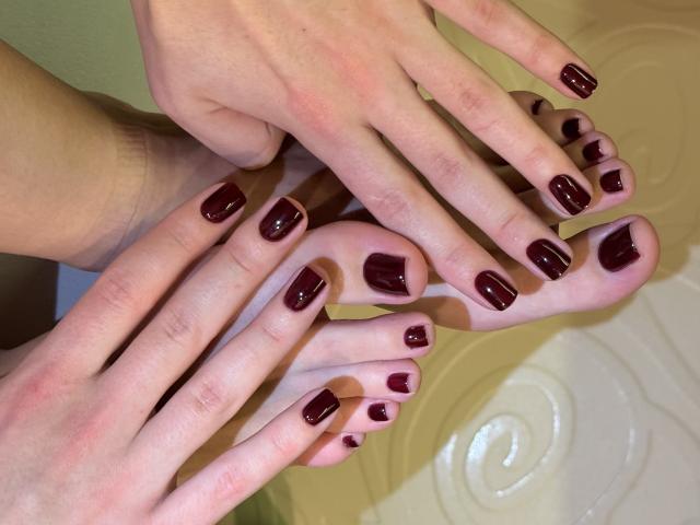 Primer plano de las manos y los pies de MargoFun, mostrando un esmalte de uñas rojo oscuro brillante en los dedos de los pies y de las manos. Los dedos extendidos sobre los dedos de los pies, creando una disposición íntima y sensual. La iluminación suave resalta la textura de la piel..