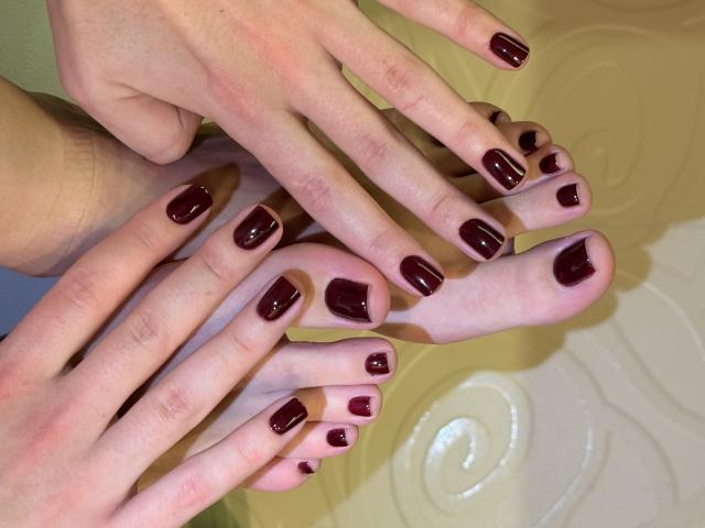 Close-up van de handen en voeten van MargoFun, waarbij netjes gemanicuurde teennagels en vingernagels te zien zijn, geverfd in een glanzend donkerrood. De handen rusten bovenop de voeten, wat een suggestieve, intieme compositie creëert..