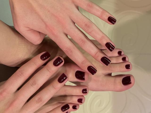 Nahaufnahme von MargoFuns Händen und Füßen, beide mit glänzendem dunkelrotem Nagellack. Die Hände liegen auf den Füßen und erzeugen so einen schichtartigen Effekt. Weiches Licht, heller Hautton. Etwas erotisch.