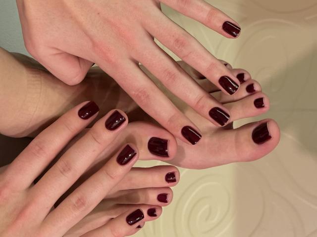 Primer plano de los pies de MargoFun, con uñas de los pies cuidadosamente manicuradas, de un rojo oscuro brillante, cruzados por las manos de MargoFun. Esmalte de uñas a juego. Las manos y los pies crean una composición sensual e íntima..