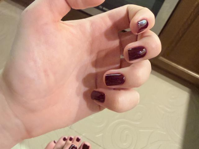 Primo piano della mano di MargoFun, con uno smalto di un intenso color borgogna scuro, e le dita leggermente incurvate. Dita dei piedi visibili sullo sfondo, colore delle unghie coordinato. Texture carnosa, illuminazione soffusa, ombre delicate. Unghie incredibilmente sexy..