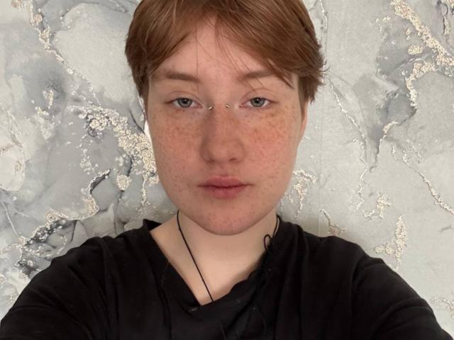 SheilahSexy heeft een lichte huid, bruin haar en sproeten. Ze draagt een zwarte blouse en kijkt recht in de camera met een neutrale uitdrukking. De achtergrond heeft een getextureerd, wit patroon. Ongelooflijk aantrekkelijk..